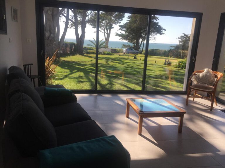 Intérieur d'un salon moderne avec canapé confortable et immense baie vitrée ouvrant sur un jardin vue mer.