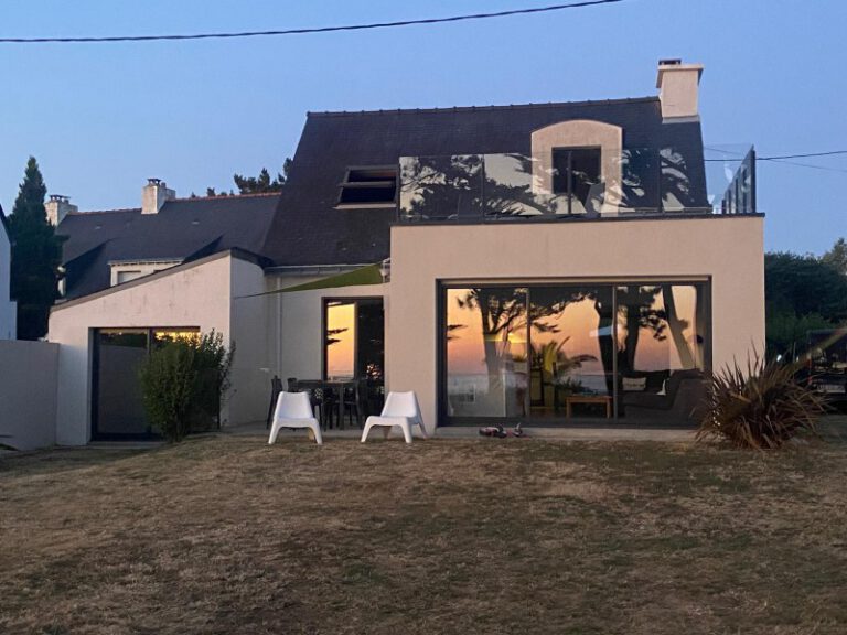 Façade d'une maison d'architecte avec grandes baies vitrées et chaises de jardin au coucher du soleil.
